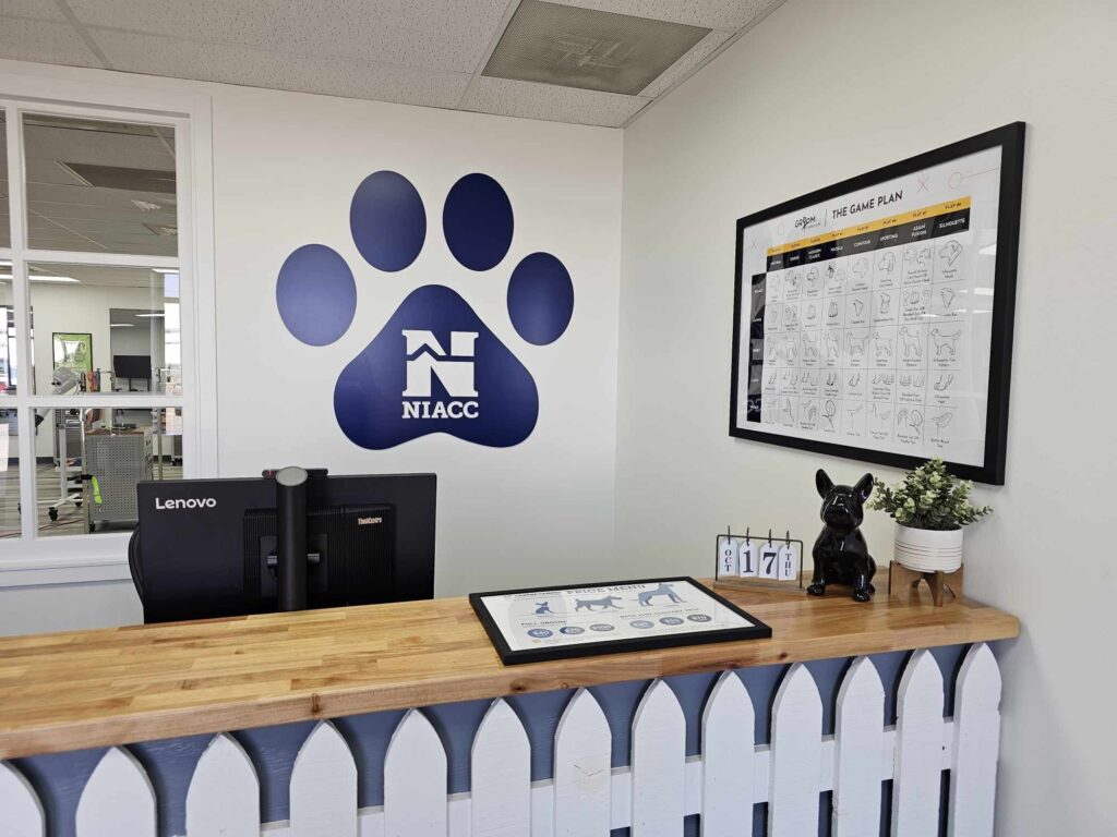 A reception desk at a pet grooming facility with a wooden countertop and white picket fence-style front. Behind the desk, a large blue paw print logo with the letters "NIACC" is displayed on the wall. On the desk, there is a computer monitor, a small black figurine of a French bulldog, a potted plant, and a decorative calendar. A framed grooming chart titled "The Game Plan" is hung on the wall, displaying different dog breed silhouettes. Through a window behind the desk, part of the grooming area is visible.