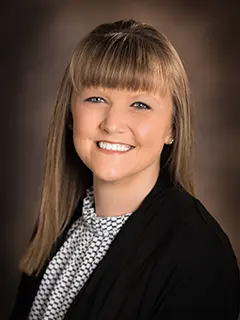 Professional headshot of Amanda Crowell, a woman with straight, light brown hair and bangs, wearing a patterned blouse and black blazer, smiling at the camera against a soft brown gradient background.