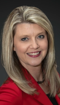 Portrait of Amy Fleming, a woman with blonde hair and blue eyes, wearing a red blazer and black top, smiling against a dark background.