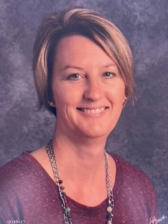 Portrait of Amy Wilhelm, a woman with short blonde hair, wearing a maroon top and a layered necklace, smiling against a textured gray background.