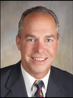 Professional headshot of Bill Feller, a man with short gray hair, wearing a dark suit, white shirt, and striped tie, smiling at the camera against a neutral gradient background.