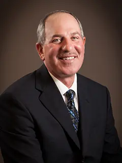 Portrait of Bill Paulus, a man wearing a dark suit, white shirt, and patterned tie, smiling against a dark brown background.