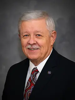 Portrait of David Steffens Jr., an older man with short gray hair and a mustache, wearing a dark suit, white shirt, and a colorful paisley tie, smiling against a dark gray background.