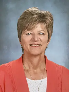 Portrait of Deb Meade, a woman with short hair wearing a coral-colored cardigan and a white top, smiling against a light blue background.