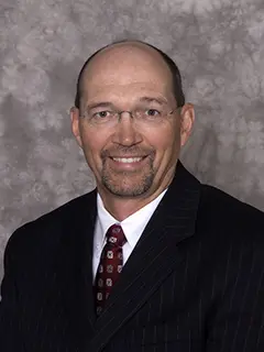 Professional headshot of Dennis Busta, a man with glasses, a bald head, and a trimmed beard, wearing a dark pinstripe suit, white shirt, and patterned tie, smiling at the camera against a textured gray background.