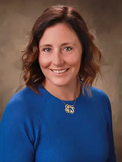 Professional headshot of Hunter Callanan, a woman with wavy, shoulder-length brown hair, wearing a blue top and a monogrammed gold necklace, smiling at the camera against a neutral brown background.