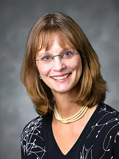 Portrait of Jean Torgeson, a woman with shoulder-length brown hair and glasses, wearing a black patterned blouse and a double-stranded pearl necklace, smiling against a gray background.