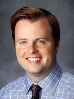 Portrait of Jeremy Peters, a man wearing a light blue checkered shirt and dark patterned tie, smiling against a gray background.