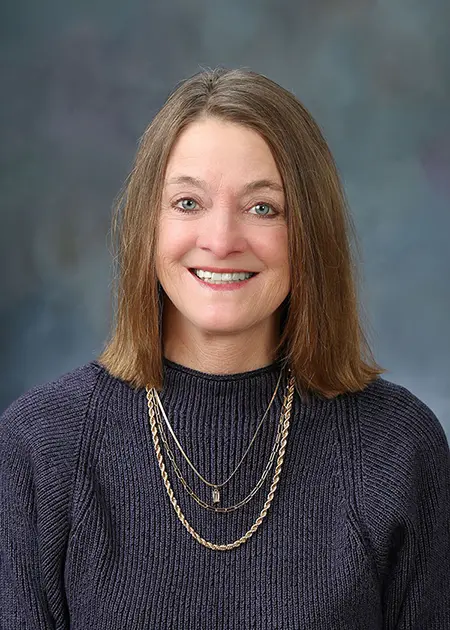 Portrait of Jodie Ferch, a woman with straight brown hair, wearing a navy sweater and layered gold necklaces, smiling in front of a neutral background.