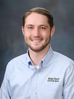 Portrait of Joe Popp, a man wearing a light blue button-up shirt with a 'Home Trust & Savings Bank' logo, smiling against a gray gradient background.