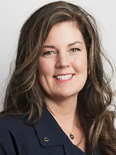 Professional headshot of Kelly Larson, a woman with long wavy brown hair, wearing a navy blouse and a gold necklace, smiling warmly at the camera against a white background.