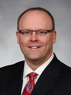 Portrait of Kirk Paulson, a man wearing glasses, a black suit, a white shirt, and a red paisley tie, smiling against a light gray background.