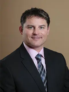 Professional headshot of Lindsey Falk, a man with short dark hair, wearing a black suit, pink shirt, and patterned tie, smiling at the camera against a plain beige background.