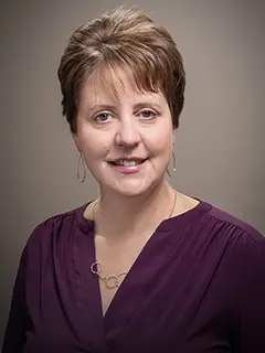 Professional headshot of Mary Amsbaugh, a woman with short brown hair wearing a purple blouse and hoop earrings, smiling at the camera against a neutral background.