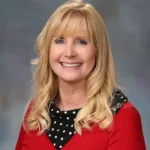 Portrait of a smiling woman with long blonde hair wearing a red blouse and a black polka dot scarf.