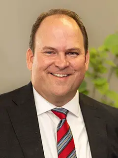 Portrait of Michael Moeller, a man wearing a black suit, white shirt, and red striped tie, smiling in front of a neutral background with green foliage.