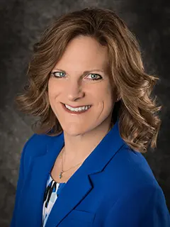 Portrait of Pamela Roberts, a woman with wavy brown hair wearing a bright blue blazer and patterned blouse, smiling against a dark gray textured background.