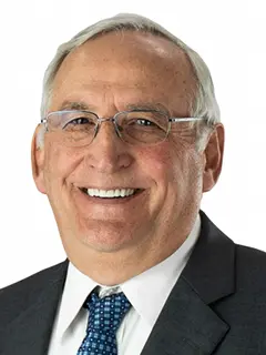 Portrait of Paul Rottinghaus, a man with short white hair wearing glasses, a dark suit, white shirt, and blue patterned tie, smiling against a white background.