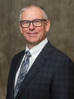 Portrait of Rod Schlader, a man with short gray hair and glasses, wearing a dark checkered suit, white shirt, and paisley-patterned tie, smiling against a neutral background.