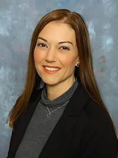 Portrait of Shanan Redinger, a woman with long brown hair wearing a dark blazer and gray turtleneck, smiling against a light blue textured background.