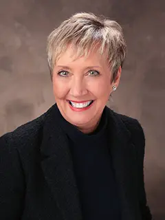 Professional headshot of Sherry Becker, a woman with short gray hair wearing a black blazer and top, smiling brightly at the camera against a softly blurred brown background.