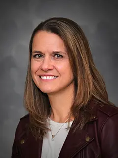 Portrait of Stephanie Nettleton, a woman with straight brown hair, wearing a burgundy jacket over a light-colored top, smiling against a gray background.