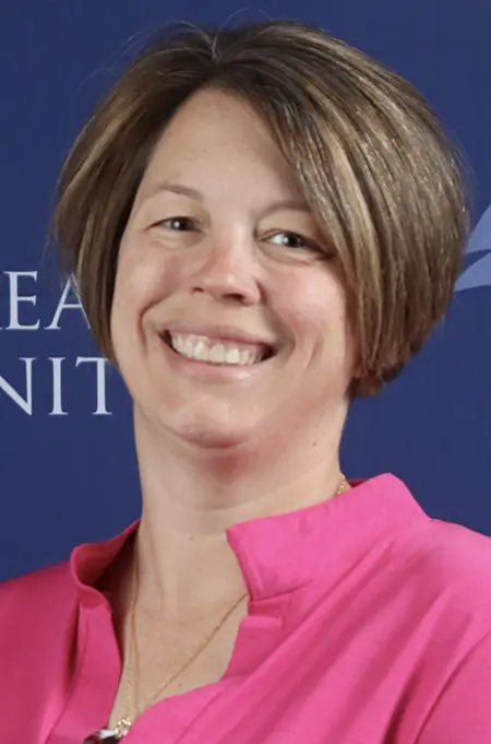 Portrait of Tanya Dadisman, a woman with short brown hair, wearing a bright pink blouse, smiling in front of a blue background with partial text visible.