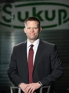 Professional portrait of Tom Mangan, a man in a dark suit and red tie, standing in front of a blurred background featuring the Sukup company logo.