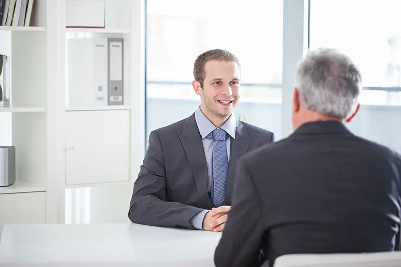 Smiling young man in a suit and tie sitting across from an older interviewer in a professional office setting.