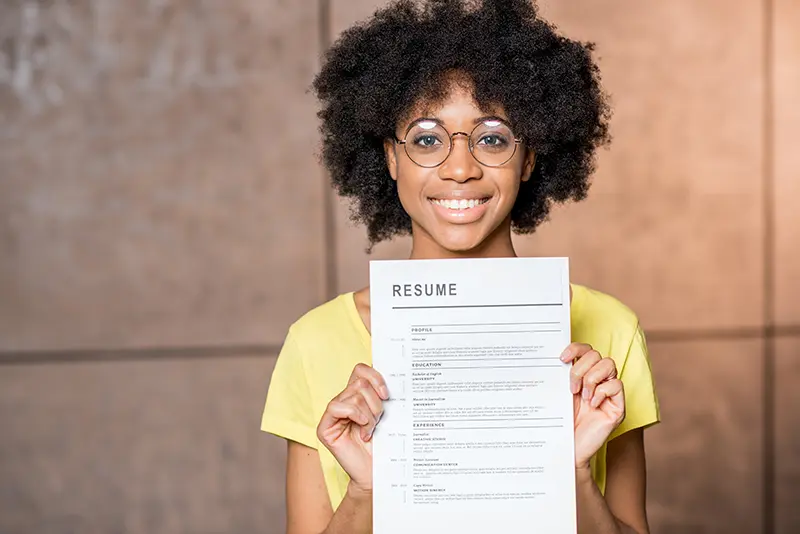 Smiling young woman with curly hair and glasses wearing a yellow shirt, holding up a printed resume in front of her.